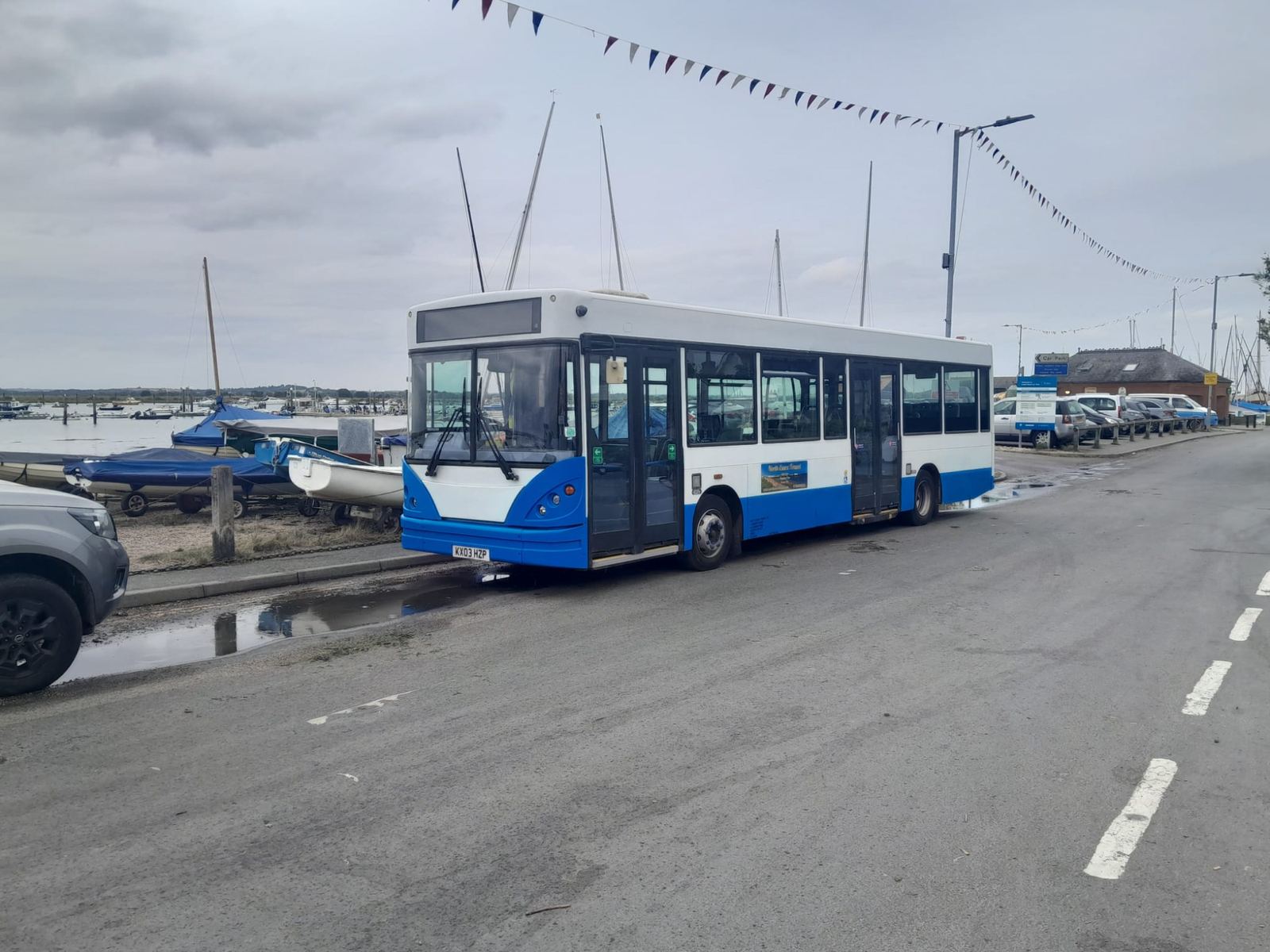 White and blue bus on a waterside road beside a harbour, with moored boats and a line of bunting