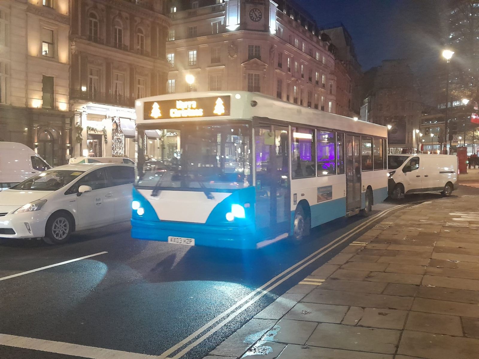 Blue and white bus with an illuminated destination display on a town street at night, with tall buildings behind